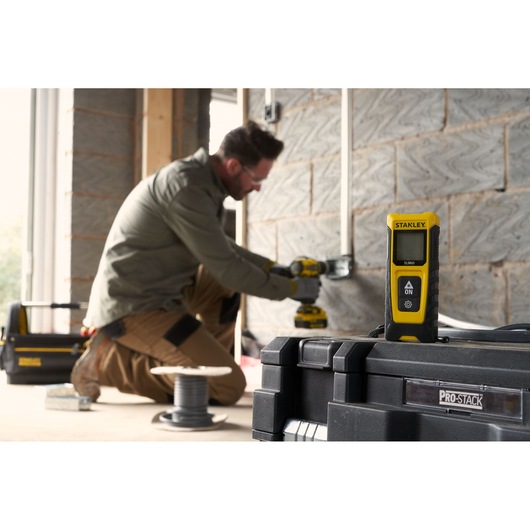Photo of STANLEY STHT77065-0 laser distance measurer on toolbox, worker using drill in background.