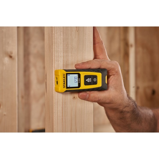 STANLEY STHT77065-0 laser distance measurer held against wooden beam, display screen lit, construction setting.