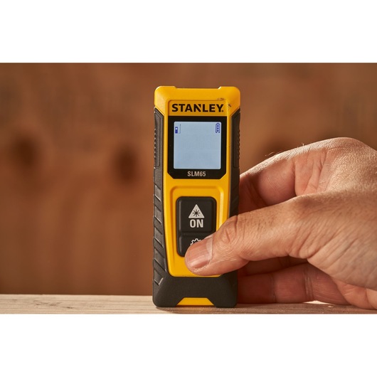 Photo of STANLEY SLM65 laser distance measurer SKU STHT77065-0 in hand, yellow and black, display screen visible.