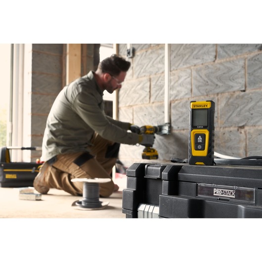 STANLEY STHT77100-0 laser distance measurer positioned on toolbox, worker installs wiring in background.
