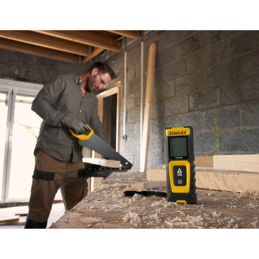 Photo of STANLEY STHT77100-0 laser distance measurer on a workshop table with sawdust, worker in background.