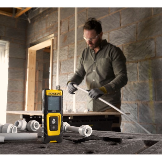 STANLEY STHT77100-0 laser distance measurer on workbench, construction materials and worker in background.