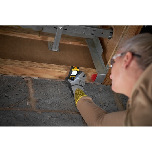 STANLEY STHT77100-0 stud finder held by gloved hand against wooden beam, product in use, indoor setting.