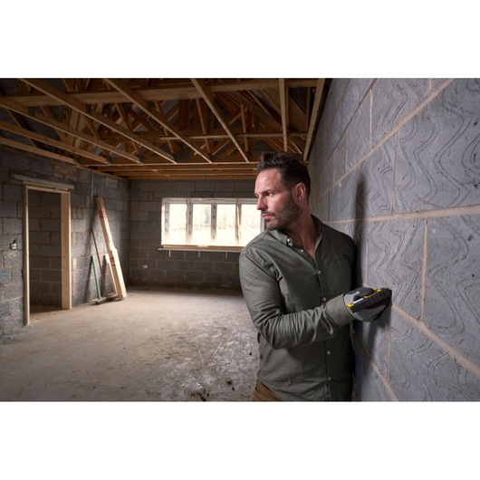 STANLEY STHT77100-0 stud sensor held by gloved hand against masonry wall in unfinished room.