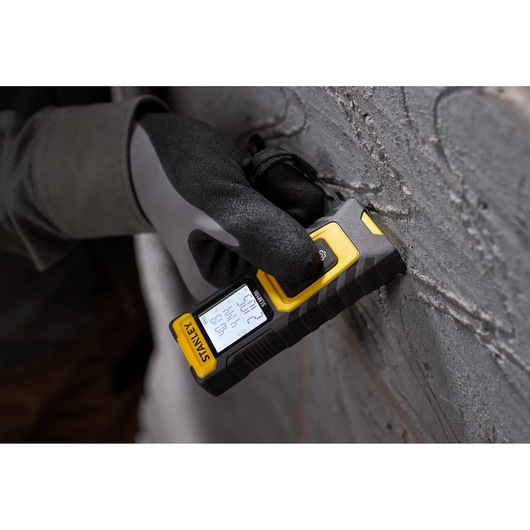 STANLEY STHT77100-0 laser distance measurer held against a concrete wall, close-up view.