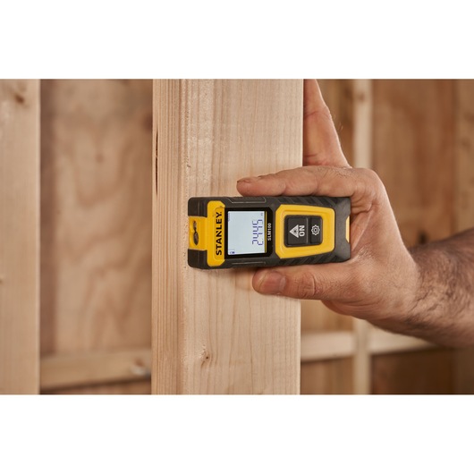 STANLEY STHT77100-0 laser distance measurer held against wood frame, display showing measurement.