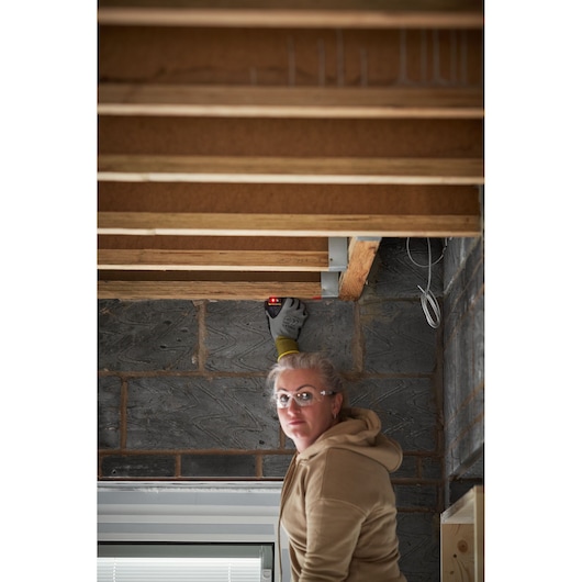 STANLEY STHT77666-0 laser measuring tool in use by a person wearing gloves, pointing at wooden beams indoors.