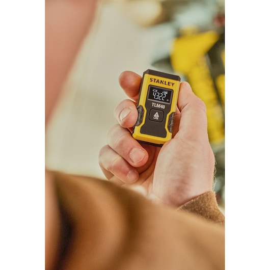 Photo of a hand holding the STANLEY STHT77666-0 digital laser distance meter, TLM40 model, showing display screen.