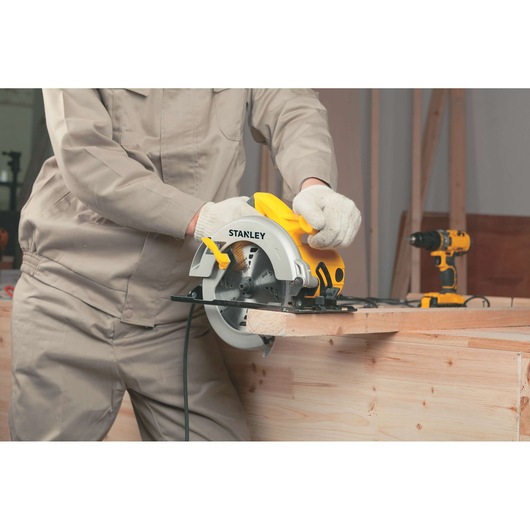 STANLEY STSC1518 circular saw in use cutting wood, worker in beige clothing and gloves, drill in background