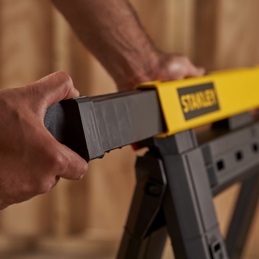 Close-up of hands adjusting STANLEY STST1-70559 black and yellow sawhorse in workshop setting.