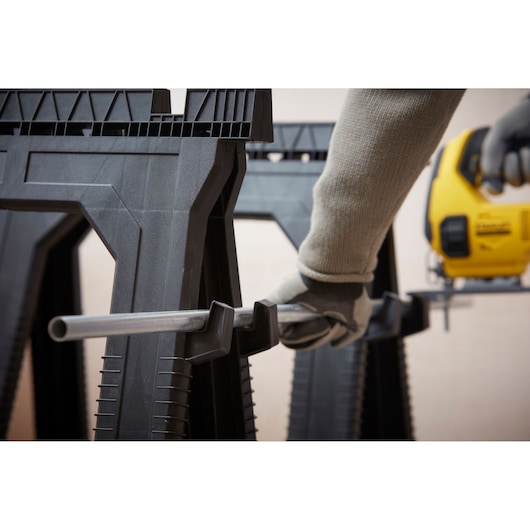 STANLEY STST1-70713 saw horse close-up with glove hand holding metal pipe, DEWALT jigsaw in background.