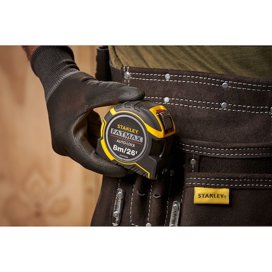 STANLEY XTHT0-33504 tape measure, black and yellow, held by gloved hand near tool belt, product side view.